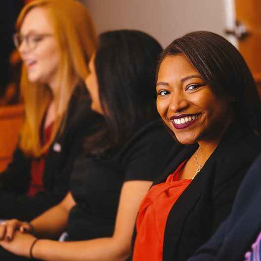 A woman in a black blazer and red top smiles at the camera while sitting among others in a row at what appears to be a professional or academic event.