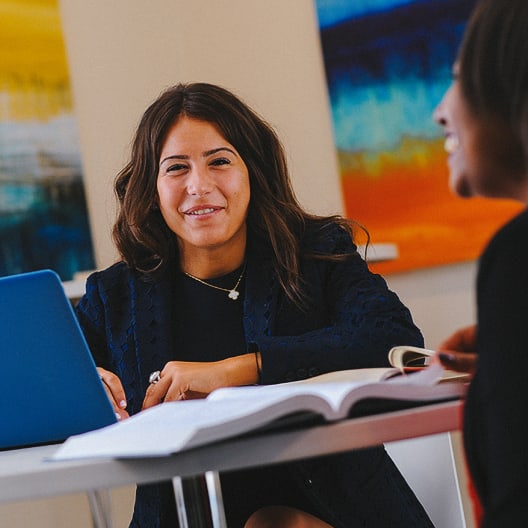 Two women sit at a table with open books and a blue laptop, smiling and talking. Colorful abstract paintings hang on the wall in the background.