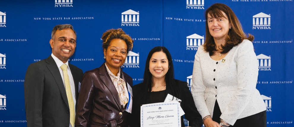 Five people stand smiling in front of a NYSBA backdrop; one woman in the center holds a certificate. The group appears to be at an award or recognition event.