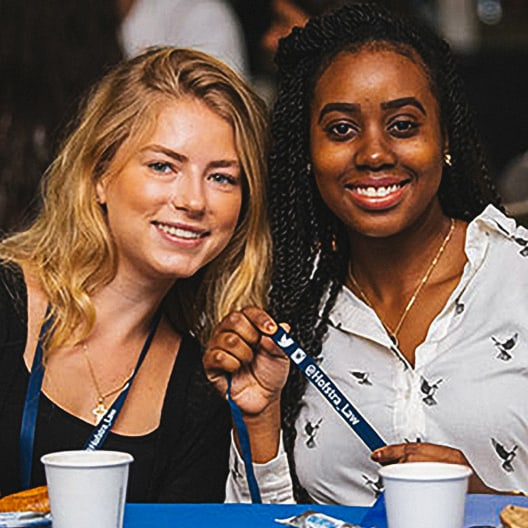 Two women smiling and sitting together at a table with white cups; one woman holds a blue lanyard with text and social media icons.