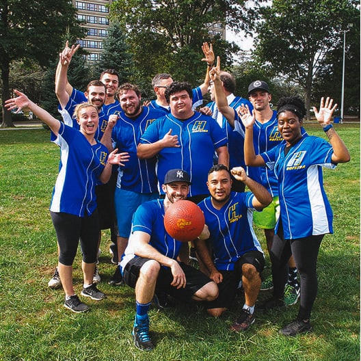 A group of people in matching blue sports jerseys pose smiling and cheering outdoors on a grassy field, with a red kickball in front of them and trees and buildings in the background.
