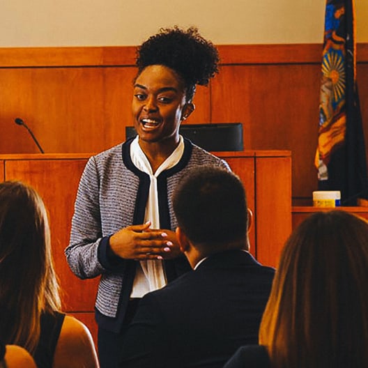 A woman stands and speaks confidently in a courtroom, addressing several seated people. A judge’s bench, microphone, and a flag are visible in the background.