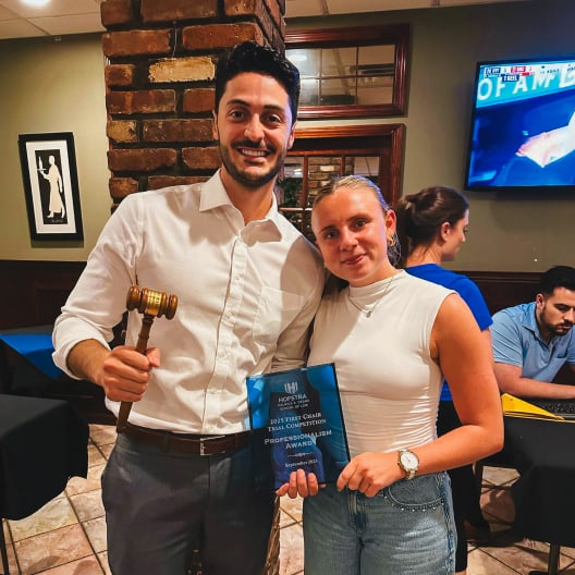 A smiling man holding a gavel and plaque stands next to a woman in a white top. They pose together indoors, with two other people working at a table in the background.