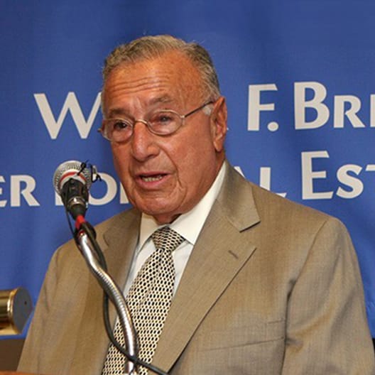 Wilbur F. Breslin in a tan suit and patterned tie speaks at a podium with a microphone, in front of a blue background with partially visible white text.