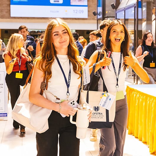 Two women with lanyards and tote bags smile while walking indoors at an event. One gives a thumbs up. Other attendees are visible in the background.