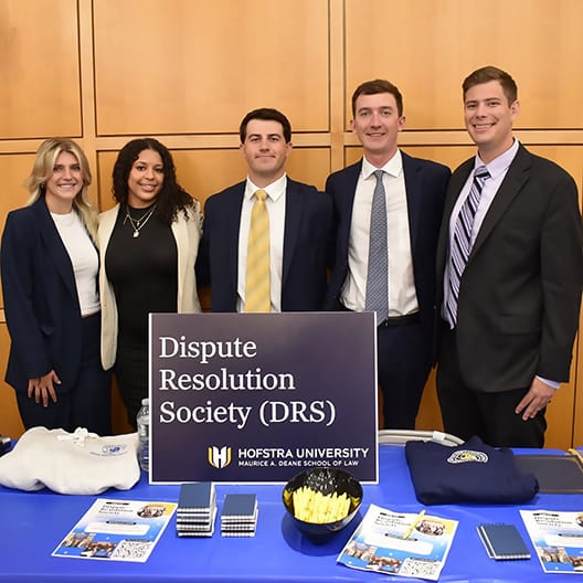 Five people in business attire stand behind a Dispute Resolution Society (DRS) table at Hofstra University School of Law, which displays brochures, snacks, and promotional items.